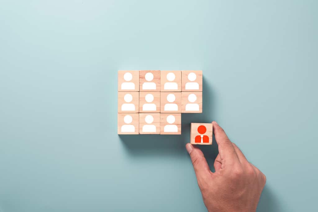 Cubos de madeira juntos, organizados em três fileiras, cada cubo tem a ilustração minimalista de uma pessoa, representando funcionários ou colaboradores. Uma mão está adicionando um último cubo com ilustração de pessoa usando roupa social na ultima fileira/coluna de cubos, para completar o cenário, simbolizando quais são os processos de RH (Recursos Humanos)