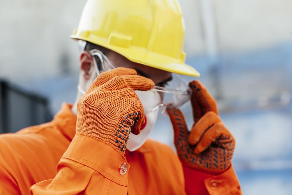 Profissional com EPIs como capacete, luvas, máscaras e colocando um óculos de proteção no rosto, simbolizando por que investir em segurança do trabalho