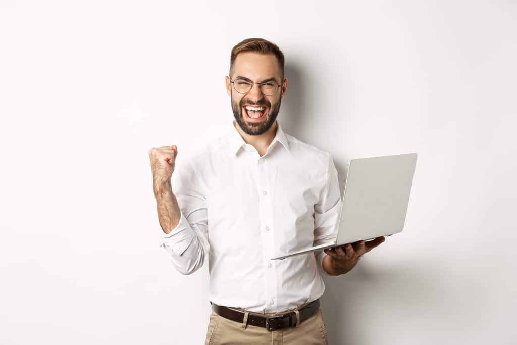 Rapaz de óculos de grau, barba e camisa social branca, sorrindo bem animado enquanto segura um notebook. Representando empresas implementando o eSocial