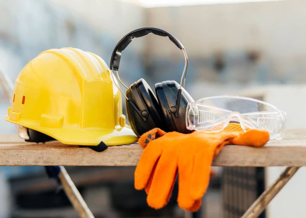 EPIs (equipamentos de proteção individual) em ambiente externo sobre uma mesa de madeira: Capacete amarelo, luvas de pano laranjas, protetores auriculares e óculos de proteção.