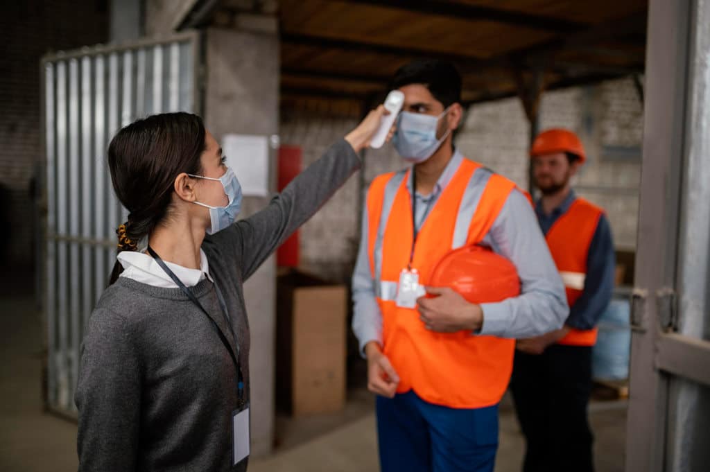 Mulher de crachá medindo temperatura de funcionário, usando um termômetro infravermelho próximo à testa dele. Ambos estão de máscara, simbolizando Saúde e Segurança do Trabalho intermitente. O funcionário está segurando um capacete, usando um colete de segurança e um crachá.