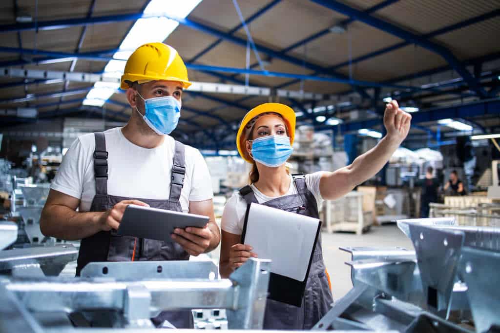 Dois profissionais com capacete, máscara e macacão, interagindo dentro de um ambiente industrial. Ela segurando uma prancheta e ele um tablet, simbolizando como fazer um mapa de risco assertivo